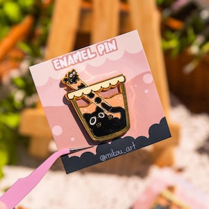 May include: A gold enamel pin featuring a cartoon illustration of a cup of boba tea with a black cat peeking out of the top. The pin is on a pink card with the text "ENAMEL PIN" and "@mikou_art".