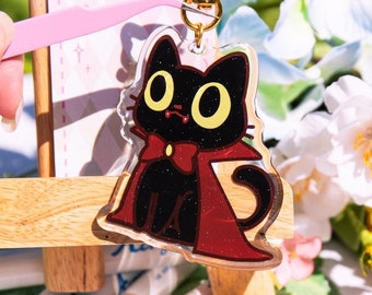 May include: A black cat with yellow eyes wearing a red cape and a gold bow tie. The cat is a keychain charm made of clear acrylic with glitter accents. The keychain is hanging from a gold chain.