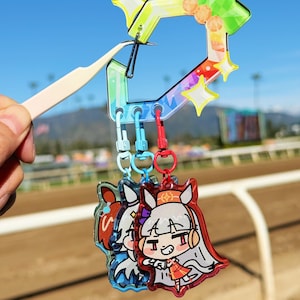 Puede incluir: Un llavero colorido con tres colgantes de personajes de estilo anime. El llavero tiene un dise&ntilde;o geom&eacute;trico de arco&iacute;ris con estrellas y detalles de gemas. Los colgantes est&aacute;n sujetos con cierres azules y rojos.