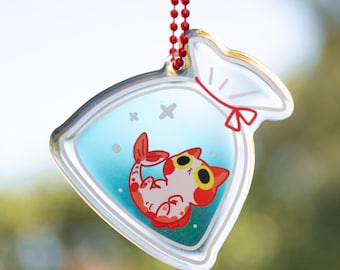 May include: A clear plastic charm with a red string attached. The charm is shaped like a bag with a red ribbon tied at the top. Inside the bag is a cartoon cat with red and white fur, swimming in blue water.