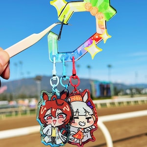 Puede incluir: Dos llaveros de personajes de estilo anime con un dije en forma de estrella de colores del arco&iacute;ris. Los llaveros presentan ilustraciones coloridas de personajes con cabello rojo y blanco. El dije es sostenido por unas pinzas contra un fondo de cielo azul.