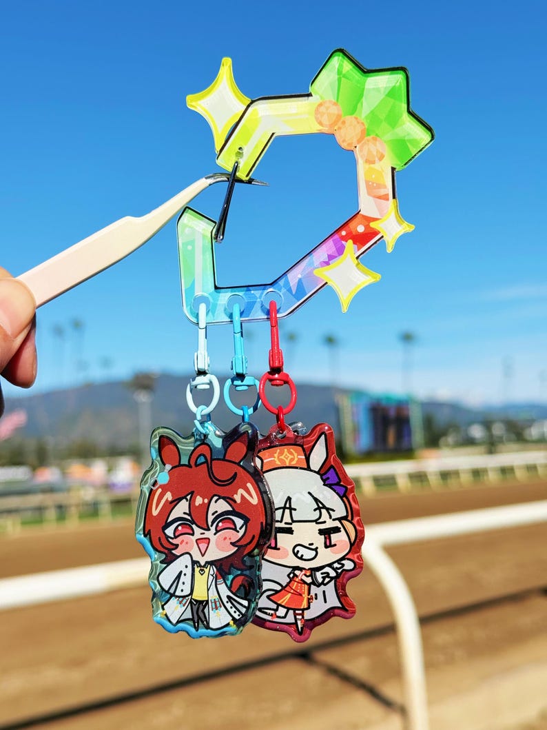 Puede incluir: Dos llaveros de personajes de estilo anime con un dije en forma de estrella de colores del arco&iacute;ris. Los llaveros presentan ilustraciones coloridas de personajes con cabello rojo y blanco. El dije es sostenido por unas pinzas contra un fondo de cielo azul.