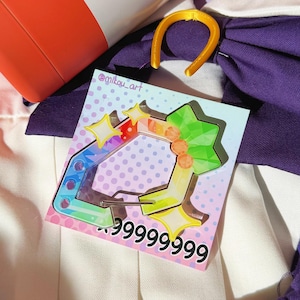 Puede incluir: Un charm de acr&iacute;lico colorido con un dise&ntilde;o de estrella y geom&eacute;trico, con el texto "999999999". El charm est&aacute; sobre un fondo de tela morada y blanca, con un objeto cil&iacute;ndrico rojo y blanco en el fondo.