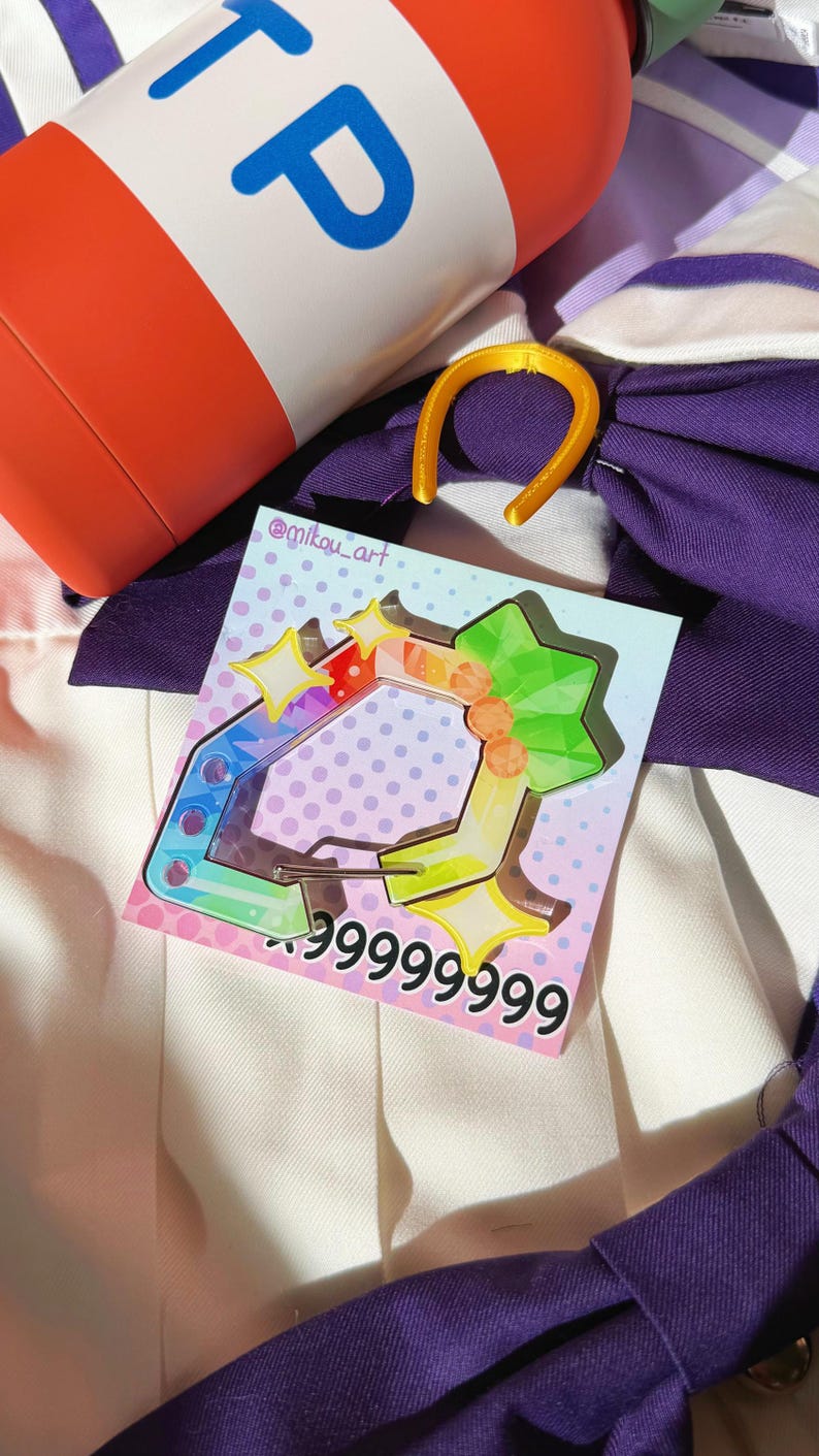 Puede incluir: Un charm de acr&iacute;lico colorido con un dise&ntilde;o de estrella y geom&eacute;trico, con el texto "999999999". El charm est&aacute; sobre un fondo de tela morada y blanca, con un objeto cil&iacute;ndrico rojo y blanco en el fondo.