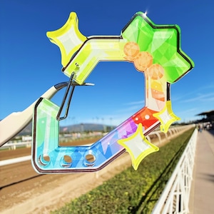 Puede incluir: Un objeto decorativo colorido en forma de estrella con un dise&ntilde;o de gradiente de arco&iacute;ris. El objeto tiene una apariencia clara y transparente con acentos de estrella amarilla y un clip de metal. El fondo muestra una pista de carreras y cielo azul.