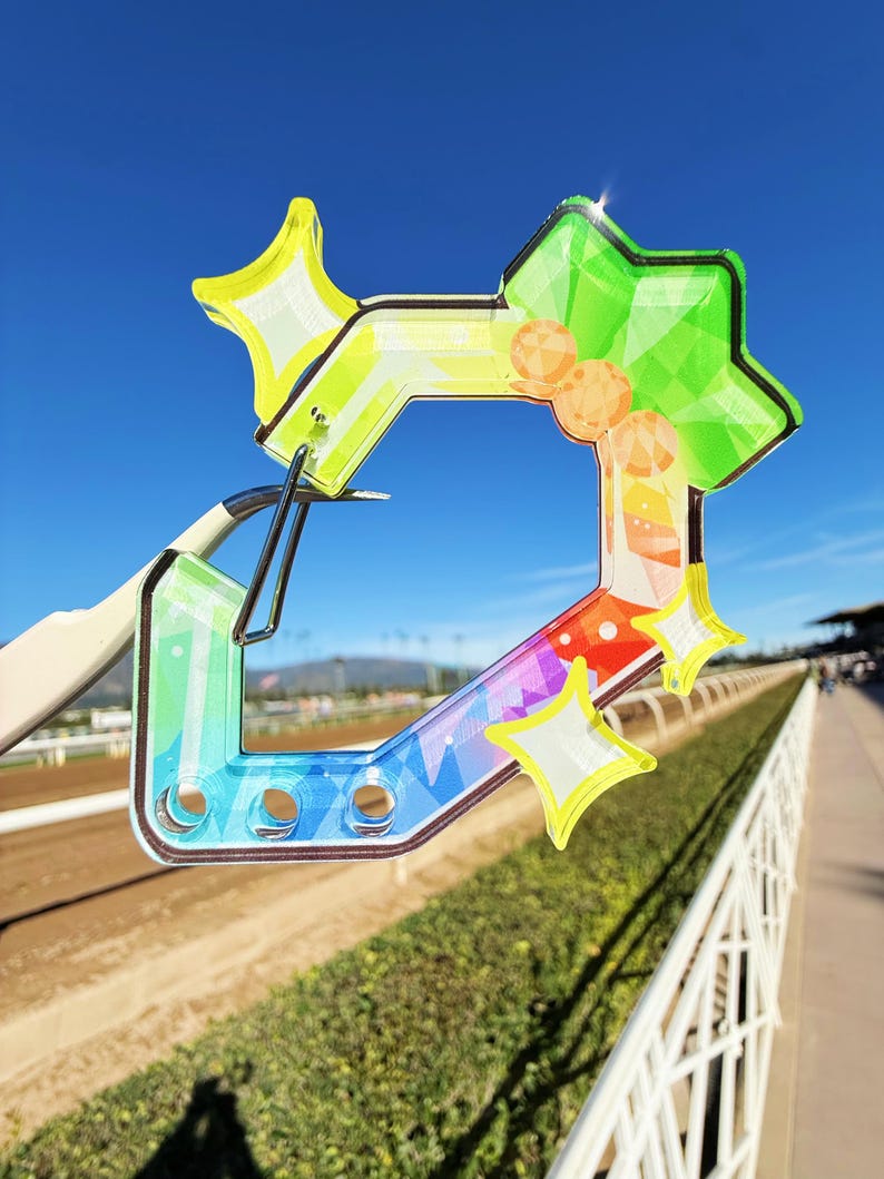 Puede incluir: Un objeto decorativo colorido en forma de estrella con un dise&ntilde;o de gradiente de arco&iacute;ris. El objeto tiene una apariencia clara y transparente con acentos de estrella amarilla y un clip de metal. El fondo muestra una pista de carreras y cielo azul.