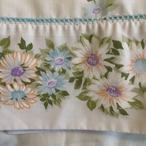 May include: White fabric with a floral pattern featuring large daisies in shades of white, orange, blue, and purple. The flowers are surrounded by green leaves. A light blue crocheted trim is at the top.