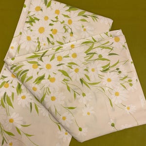 May include: Cream-colored fabric with a repeating pattern of white daisies with yellow centers and green leaves. The fabric is folded, showing multiple layers. The background is a solid olive green.