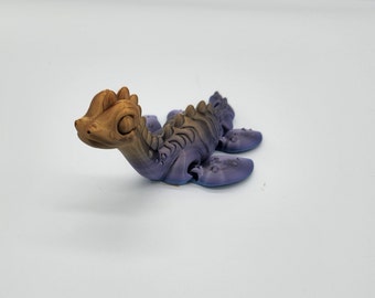 May include: A small, 3D printed, purple and gold dinosaur figurine with a long neck and tail. The dinosaur has a friendly face with large eyes and a small mouth.