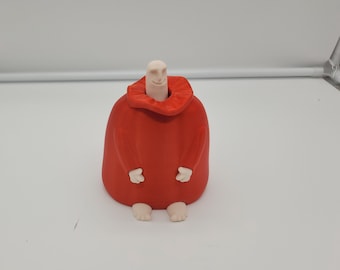 Peut inclure: Une figurine rouge fantaisiste avec une tête souriante de couleur pâle et de petites mains et pieds blancs. La figurine porte une grande cape rouge arrondie. Le design est simple et cartoon, avec une finition lisse et mate.
