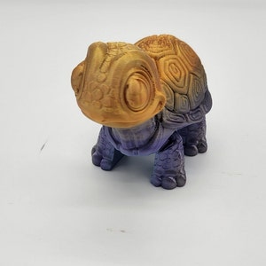 May include: A small turtle figurine featuring a gradient color transition from a metallic gold head and shell top to a deep purple on the lower shell and legs. The turtle has large, prominent eyes and a textured, patterned shell surface.