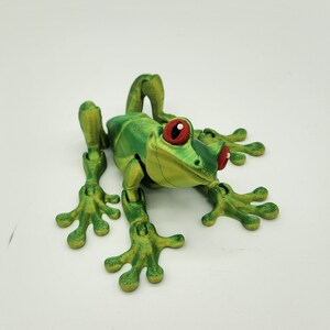 May include: A green and yellow articulated frog toy with red eyes. The frog has a textured surface and is posed in a sitting position with its legs outstretched.