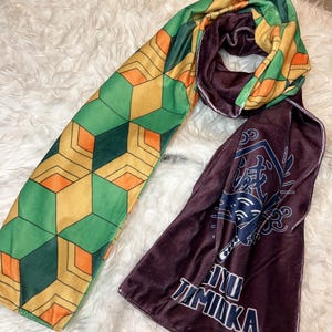 May include: A long scarf with two distinct patterned sections. One side features a geometric design in green, yellow, and orange. The other side is a deep burgundy with Japanese text and a graphic. The scarf is made of a soft, plush material.