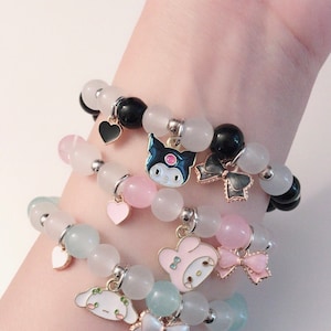 Op de afbeelding: Drie gelaagde armbanden met pastelkleurige kralen en bedels. De bovenste armband heeft zwarte kralen en een zwarte hartbedel. De middelste armband heeft roze kralen en een roze hartbedel. De onderste armband heeft lichtblauwe kralen en een witte strikbedel. Elke armband heeft een andere cartoonfiguurbedel.