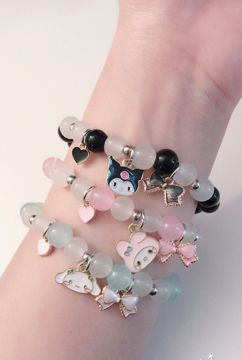 Op de afbeelding: Drie gelaagde armbanden met pastelkleurige kralen en bedels. De bovenste armband heeft zwarte kralen en een zwarte hartbedel. De middelste armband heeft roze kralen en een roze hartbedel. De onderste armband heeft lichtblauwe kralen en een witte strikbedel. Elke armband heeft een andere cartoonfiguurbedel.