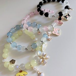 Op de afbeelding: Vier armbanden met verschillende gekleurde kralen en bedels. De armbanden zijn gemaakt van lichtblauwe, roze, gele en zwarte kralen. De bedels omvatten een hart, een strik, een bloem, een hond, een konijn en een kat.