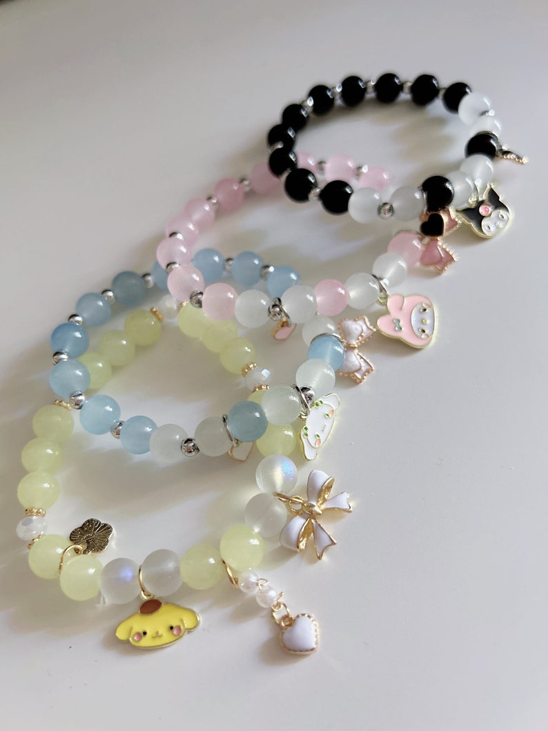 Op de afbeelding: Vier armbanden met verschillende gekleurde kralen en bedels. De armbanden zijn gemaakt van lichtblauwe, roze, gele en zwarte kralen. De bedels omvatten een hart, een strik, een bloem, een hond, een konijn en een kat.