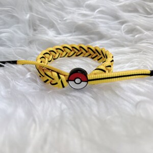 May include: Yellow and black braided bracelet with a red, white, and black Pokeball charm. The bracelet has a black tag with the text "ACG" on it.