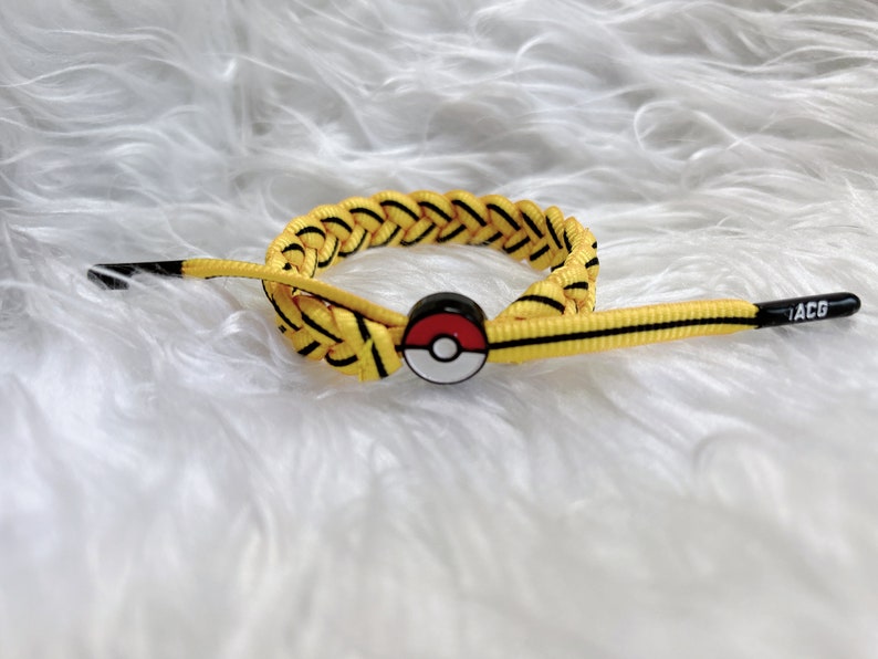 May include: Yellow and black braided bracelet with a red, white, and black Pokeball charm. The bracelet has a black tag with the text "ACG" on it.