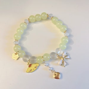 Op de afbeelding: Een lichtgroene kralenarmband met een gouden bedel van een cartoonhond, een witte strikbedel en een gouden hartbedel. De armband heeft ook een klein gouden label met de tekst "Hana smile".