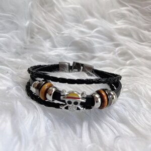 May include: A black leather bracelet with a silver skull charm and a yellow and red hat on top. The bracelet has brown wooden beads.