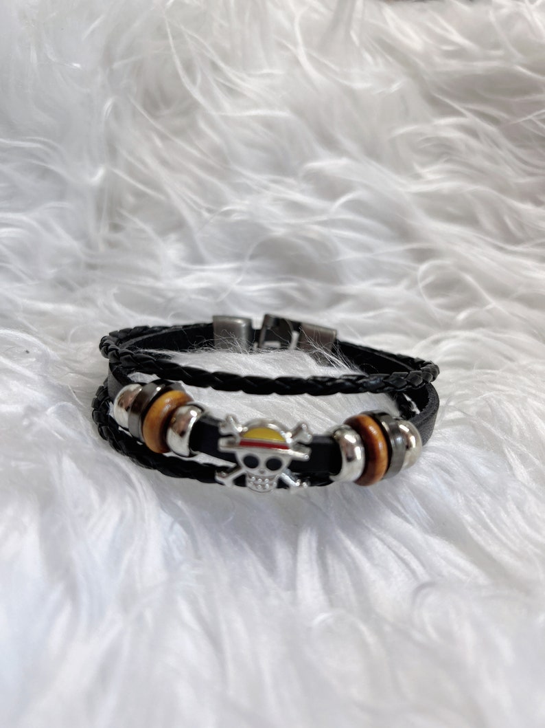 May include: A black leather bracelet with a silver skull charm and a yellow and red hat on top. The bracelet has brown wooden beads.