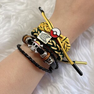 May include: A set of four bracelets featuring a black leather bracelet with a skull charm, a yellow and green braided bracelet with a Pokemon charm, a yellow and green braided bracelet with a winged symbol, and a yellow and green braided bracelet with a gold-colored charm.