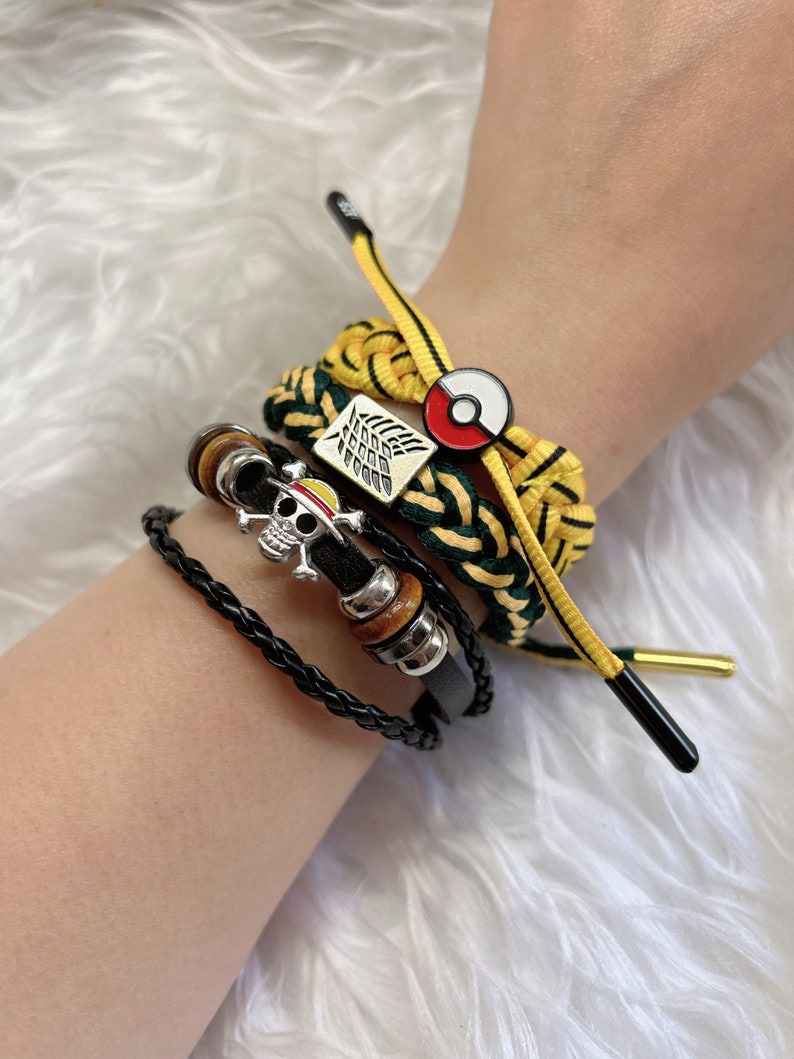 May include: A set of four bracelets featuring a black leather bracelet with a skull charm, a yellow and green braided bracelet with a Pokemon charm, a yellow and green braided bracelet with a winged symbol, and a yellow and green braided bracelet with a gold-colored charm.