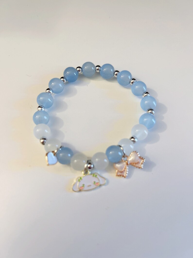 Op de afbeelding: Een lichtblauwe en witte kralen armband met een bedel van een cartoon hond en twee strikken.