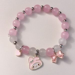 Op de afbeelding: Een roze en witte kralen armband met een bedel van een roze cartoonfiguur met een strik, een roze hartbedel en een roze strikbedel.