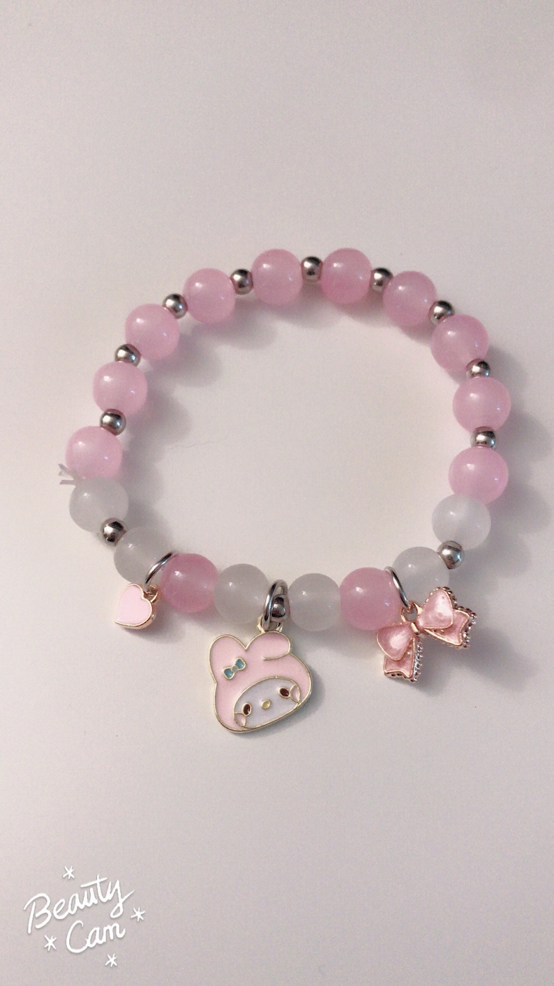 Op de afbeelding: Een roze en witte kralen armband met een bedel van een roze cartoonfiguur met een strik, een roze hartbedel en een roze strikbedel.