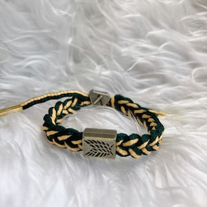 May include: A green and gold braided bracelet with a silver charm featuring a leaf design.