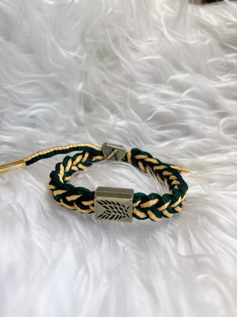 May include: A green and gold braided bracelet with a silver charm featuring a leaf design.