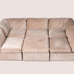 May include: A modular sectional sofa upholstered in a light beige velvet fabric. The sofa features a low-profile design with three distinct seating sections, each displaying a subtle diagonal quilted pattern across the cushions and backrests.