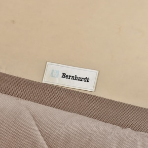 May include: A beige fabric upholstery surface featuring a rectangular white label with a blue logo and the text Bernhardt in black lettering. The fabric has a visible woven texture and is stitched to a smooth, light-colored panel.