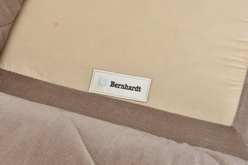 May include: A beige fabric upholstery surface featuring a rectangular white label with a blue logo and the text Bernhardt in black lettering. The fabric has a visible woven texture and is stitched to a smooth, light-colored panel.