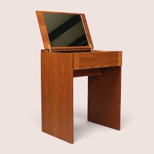 May include: A wooden vanity table featuring a warm brown wood grain finish, a rectangular flip-top mirror attached with brass hinges, and a single front drawer. The structure is supported by two side panels, creating an open space underneath.