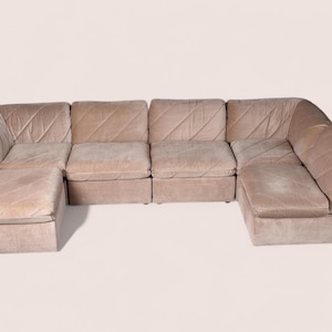 May include: A modular sectional sofa in a light beige velvet fabric featuring a low-profile design with multiple seating sections and a matching ottoman. The upholstery displays a subtle diagonal stitched pattern across the back and seat cushions.