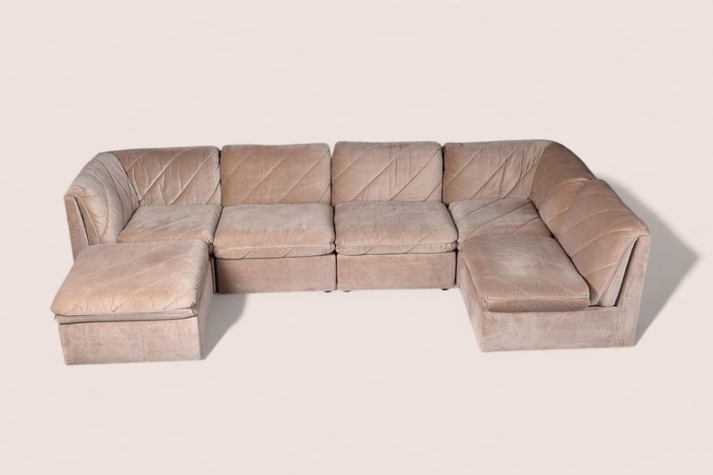 May include: A modular sectional sofa in a light beige velvet fabric featuring a low-profile design with multiple seating sections and a matching ottoman. The upholstery displays a subtle diagonal stitched pattern across the back and seat cushions.