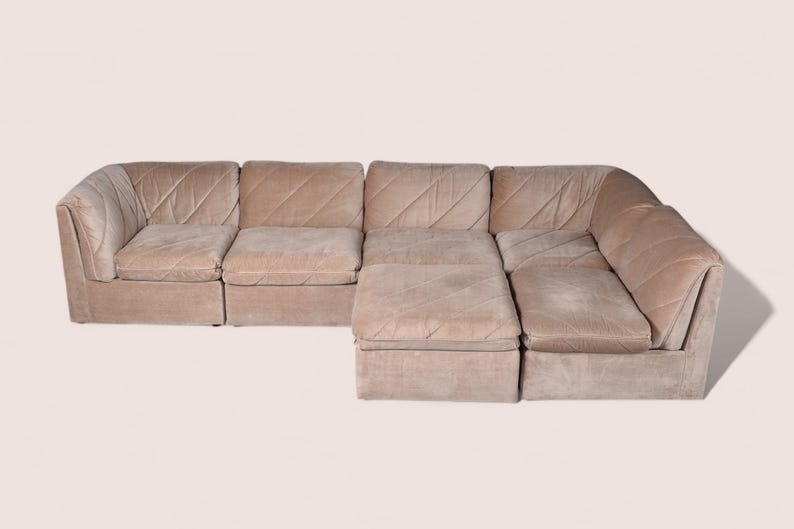 May include: A modular sectional sofa upholstered in a light beige fabric with a diagonal quilted pattern. The sofa consists of multiple connected seating units, including a corner piece and an extended chaise section, featuring low backrests.