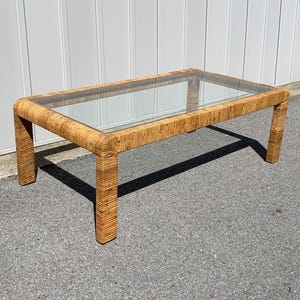 May include: A rectangular wicker coffee table with a glass top. The table has a natural brown color and is made of woven wicker.