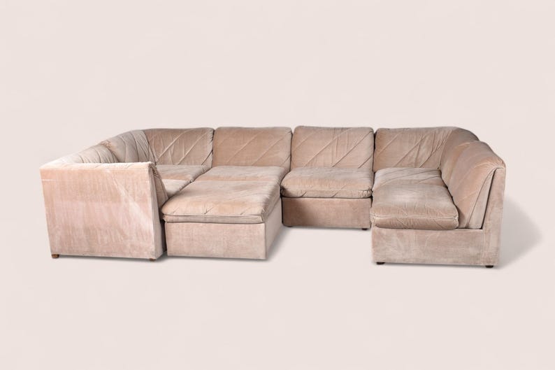May include: A modular sectional sofa upholstered in a light beige fabric. The sofa features multiple connected seating sections with low backrests and a distinct diagonal stitched pattern across the cushions, creating a unified seating arrangement.