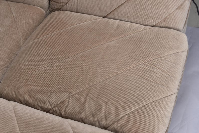 May include: A beige sofa cushion featuring a textured fabric surface with a prominent diagonal stitched pattern. The cushion is square-shaped with visible seams that create a geometric grid across the upholstery, showcasing a neutral tone.
