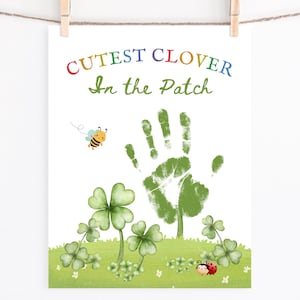 以下が含まれることがあります： クローバーの形をした緑色の手形が印刷されたアートプリント。緑色のクローバーとミツバチに囲まれています。プリントの上部には「Cutest Clover In the Patch」というテキストが虹色で書かれています。