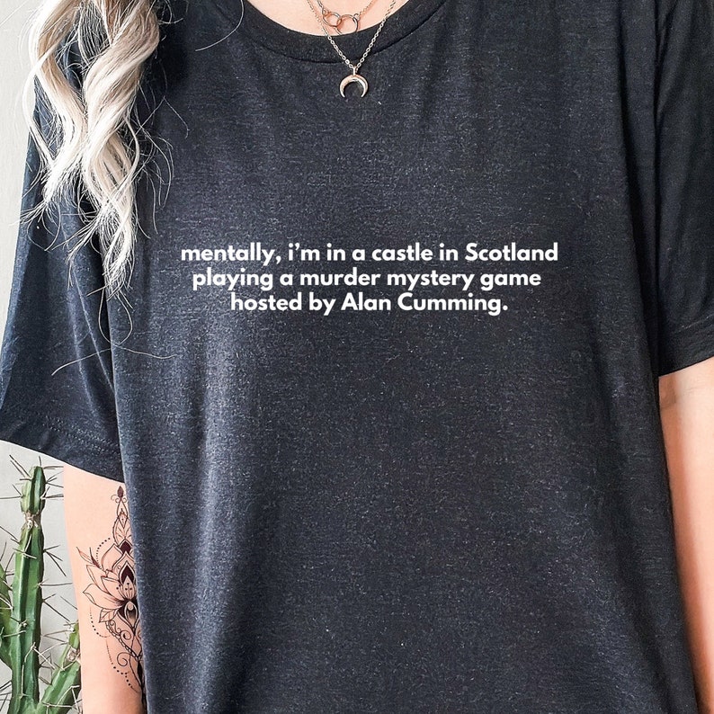 May include: A black t-shirt with white text that reads "mentally, i'm in a castle in Scotland playing a murder mystery game hosted by Alan Cumming."
