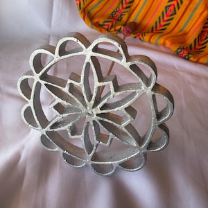 May include: A silver-colored rosette iron with a floral design, resting on a white surface. The iron has a handle and a decorative pattern. In the background, a yellow fabric with colorful stripes and geometric patterns is visible.