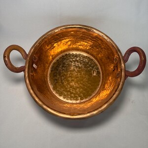 May include: A hammered copper bowl with two loop handles. The bowl has a shiny, reflective interior and a textured exterior. The handles are a darker shade of copper. The bowl is round and appears to be handmade.