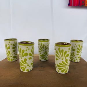 May include: Five ceramic shot glasses with a cream-colored base and a vibrant green floral design. The rims and interiors are a dark brown. The glasses are arranged on a wooden surface.