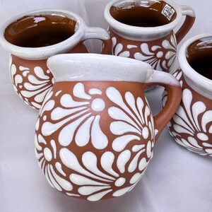 May include: Set of four ceramic mugs. Each mug has a brown base with white floral designs and a white rim. The handles and interior are brown. The mugs are arranged on a white surface.