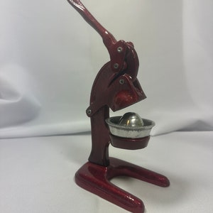 May include: A red metal citrus press with a lever and a silver bowl. The press is designed to squeeze juice from citrus fruits.